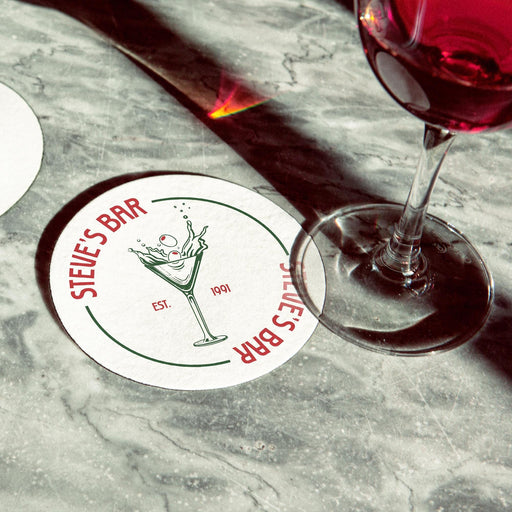 Personalized dirty martini coaster placed beside cocktail glasses on a marble bar surface, featuring the custom bar name, martini illustration, and established year.
