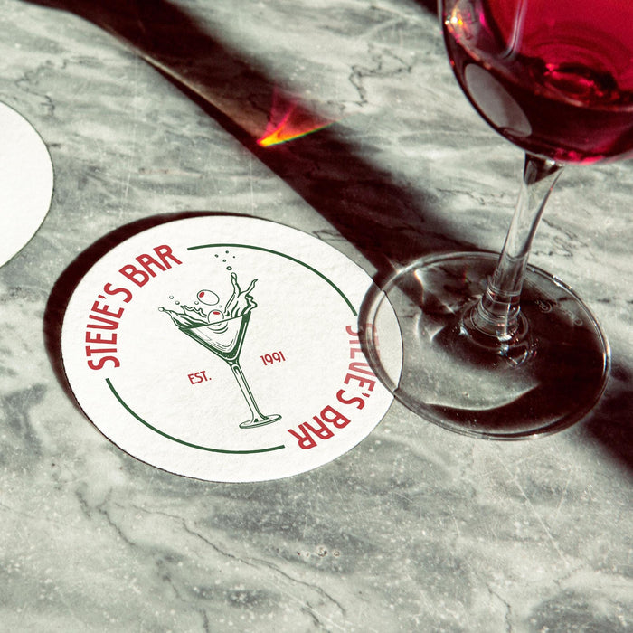 Personalized dirty martini coaster placed beside cocktail glasses on a marble bar surface, featuring the custom bar name, martini illustration, and established year.