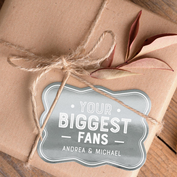 Personalized “Your Biggest Fans” gift tag attached to a wrapped kraft paper gift using natural twine. The metallic silver foil finish creates an elegant keepsake-style wedding tag.