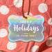 Personalized Happy Holidays rounded rectangle hang tag displayed against red and white polka dot holiday wrapping paper.