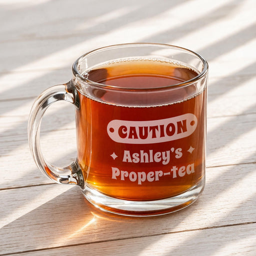 A glass mug filled with tea, placed on a wooden surface, with a graphic engraved on it, which says "Caution Ashley's Proper-tea"
