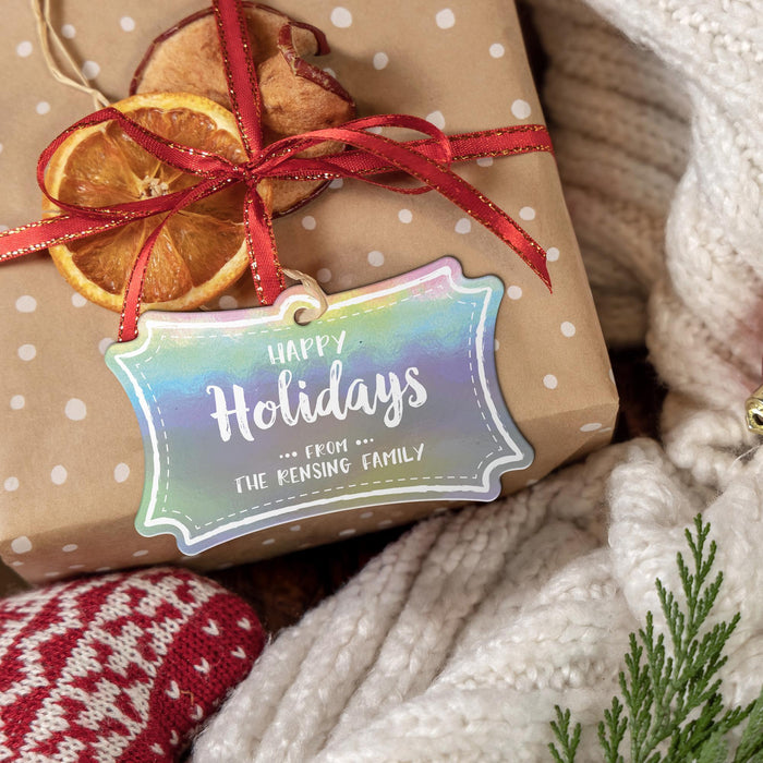 Personalized Happy Holidays hang tag tied to a kraft paper gift with red ribbon, dried orange slices, greenery, and a cozy knit background.