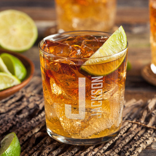 Personalized whiskey glass displayed with whiskey, ice, and fresh lime on a rustic wooden surface. The large initial and vertical name engraving is clearly visible, showing how the finished custom glass looks in a home bar setting.