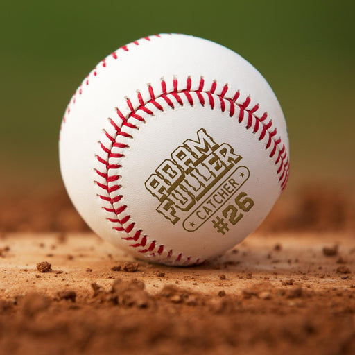 A personalized baseball with red stitching, featuring the name "Adam Fuller" and the number "#26" engraved on it, resting on a dirt surface.