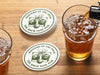 Custom round pulpboard coasters are shown sitting on a wooden table next to two drink glasses. The custom coasters feature a golf ball centered between two whiskey glasses clinking together and splashing out with customizable text wrapping around the illustration. The coaster design is printed in olive green.