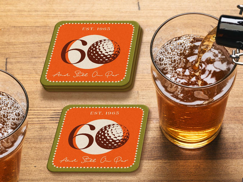 Custom square pulpboard coasters are shown laid out on a wooden surface. Two beer mugs are shown next to the coasters. The coaster features design with illustrated golf ball, custom age and year, and vivid orange, cream, and green background.