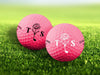 Two golf balls with the letters 'TS' and a floral golf club design printed on them, placed on a grassy field.