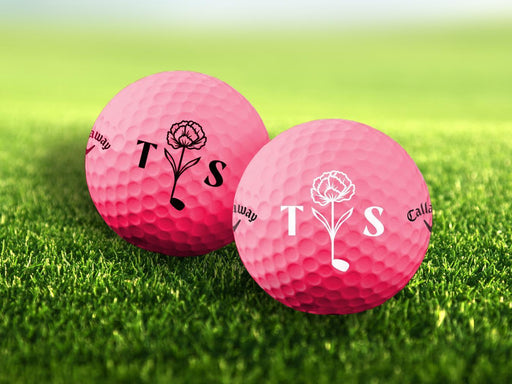 Two golf balls with the letters 'TS' and a floral golf club design printed on them, placed on a grassy field.