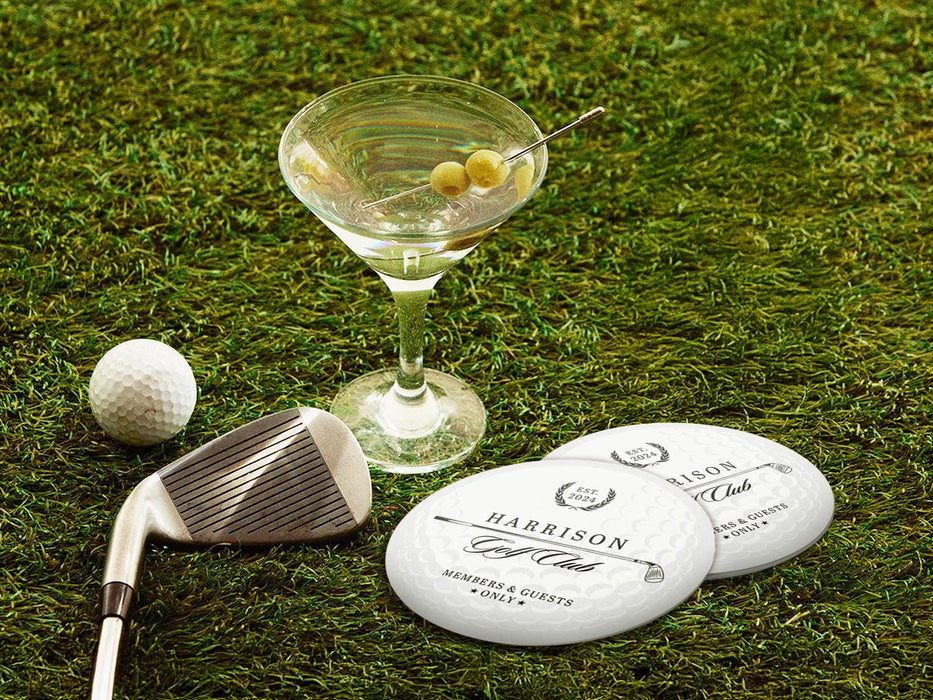 Two custom coasters sit on a grassy golf course next two a martini glass, golf club, and golf ball. The coasters feature a golf club and overlayed golf ball design with the text Members & Guests Only between stars. The last name, club name, and established year can be customized.