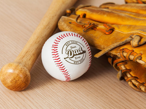 Baseball, wooden bat, and glove sits on wooden surface. Baseball has Best Dad Ever design engraved on it.