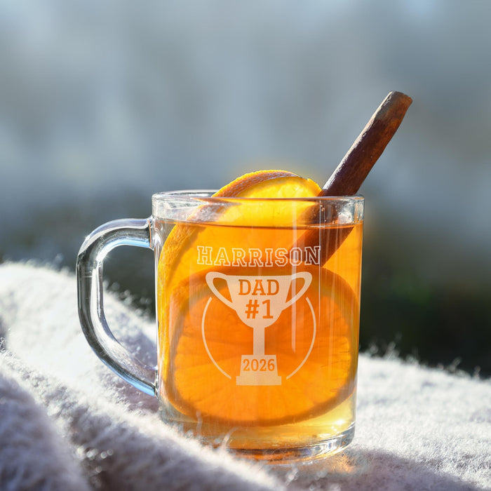 A glass mug filled with a yellow liquid, likely tea, and garnished with an orange slice and a cinnamon stick. The mug has a personalized message engraved on it, which reads "HARRISON Dad #1 2026".
