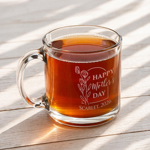A glass mug filled with a warm beverage, likely tea, sits on a wooden surface. The mug has a personalized message engraved on it, celebrating Mother's Day.