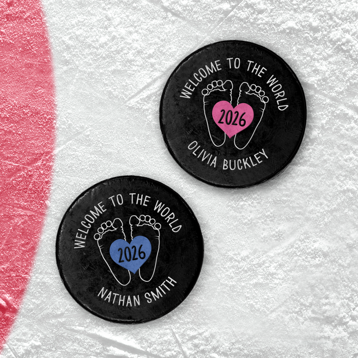 Two personalized hockey pucks with a UV printed baby birth announcement design featuring baby feet, a heart with the birth year, and custom names.