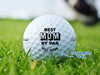 A golf ball with the text "BEST MOM BY PAR EST. 2026" printed on it, placed on a grassy field.