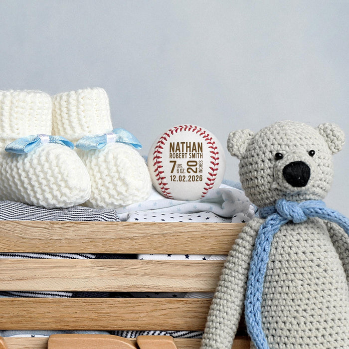 Personalized engraved baseball birth announcement shown with blue baby booties, a knitted teddy bear, and nursery decor.