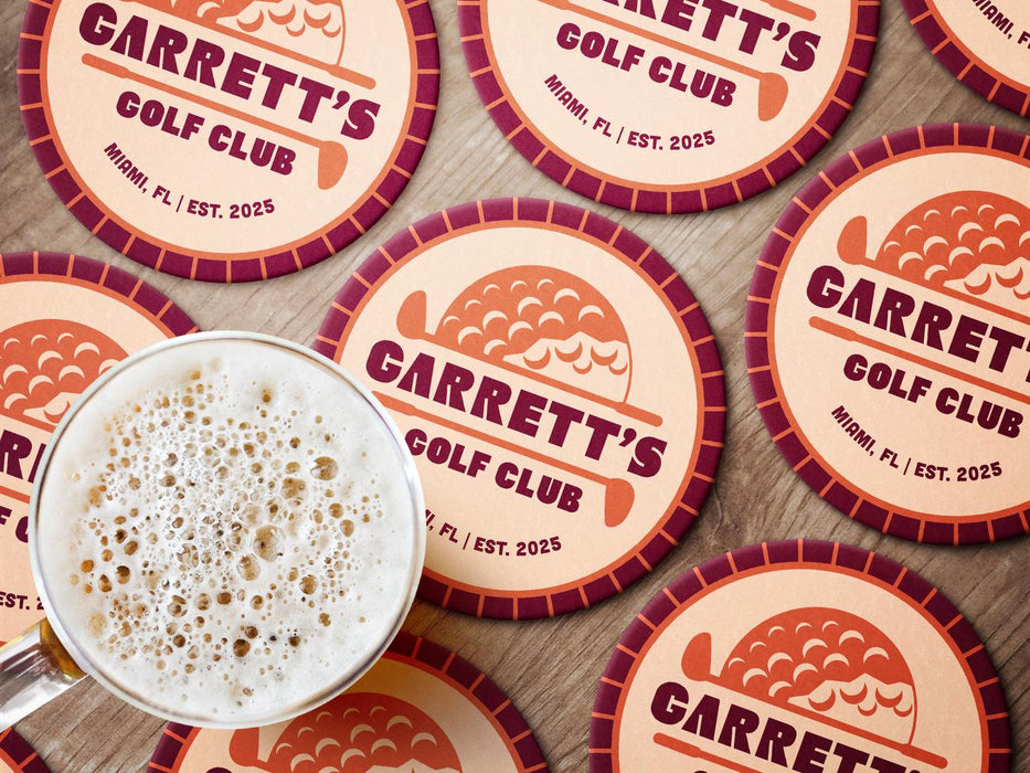 Custom round pulpboard coasters are shown sitting on a wooden surface with a beer mug on top of one. The custom coasters feature design printed with bold golf imagery, striped radial border, and custom text.