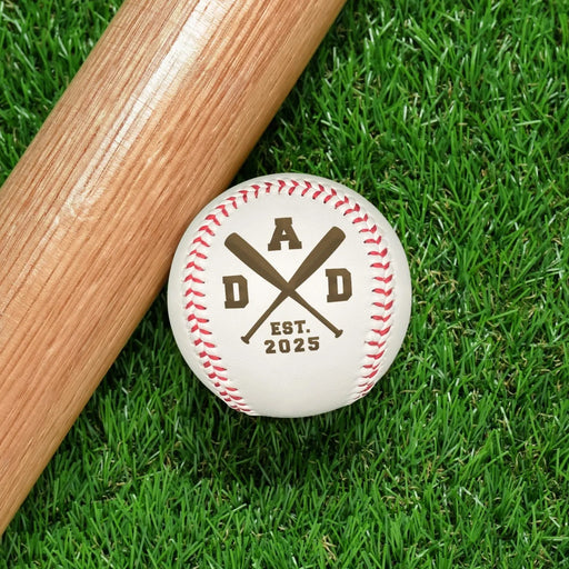 Baseball sits on grass next to baseball bat. Baseball has DAD design engraved on it.