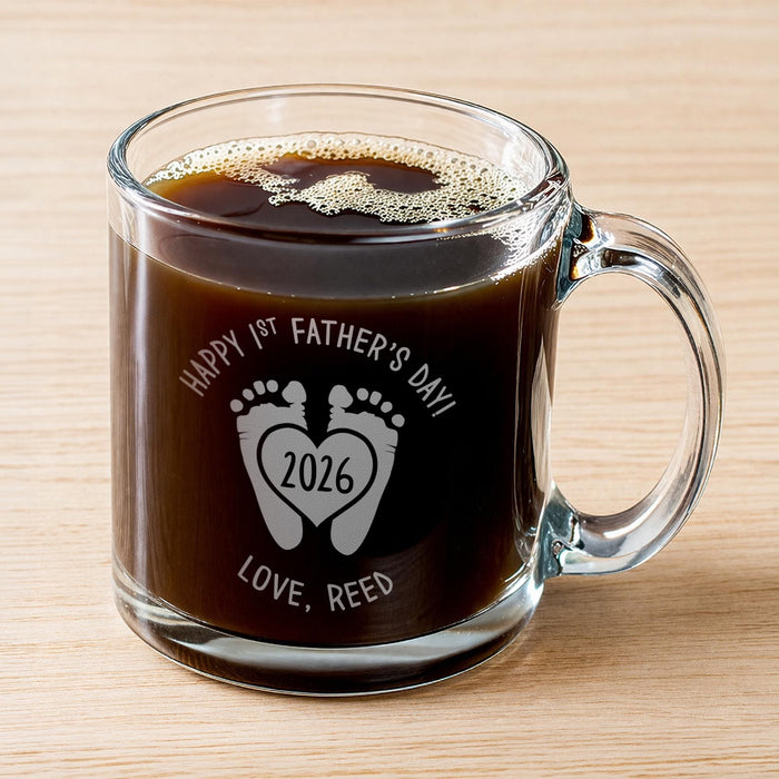 A glass mug filled with a dark liquid, likely coffee, with foam on top. The mug has an engraved message that reads "Happy 1st Father's Day 2026 Love, Reed".