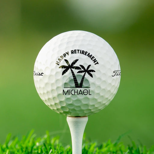 Personalized retirement golf ball featuring the Happy Retirement with palm trees design with a custom name. Displayed on a golf tee against a golf course background.