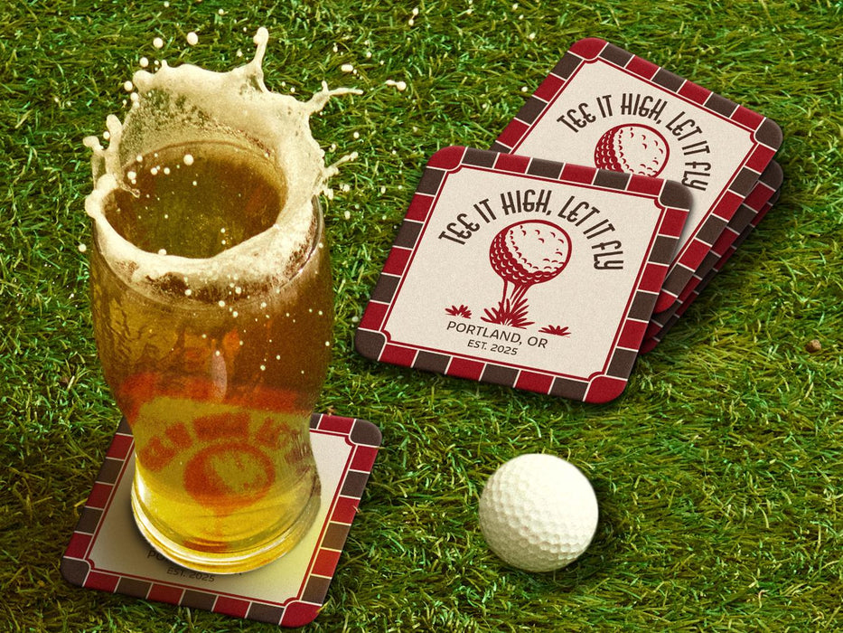Custom square pulpboard coasters are shown sitting on a grassy, green golf course next to a pint of beer and a white golf ball. The pint of beer has a golf ball splashing into it and has a coaster underneath it. The coasters feature design with an illustration of a golf ball on a tee, checkerboard border, and custom text.