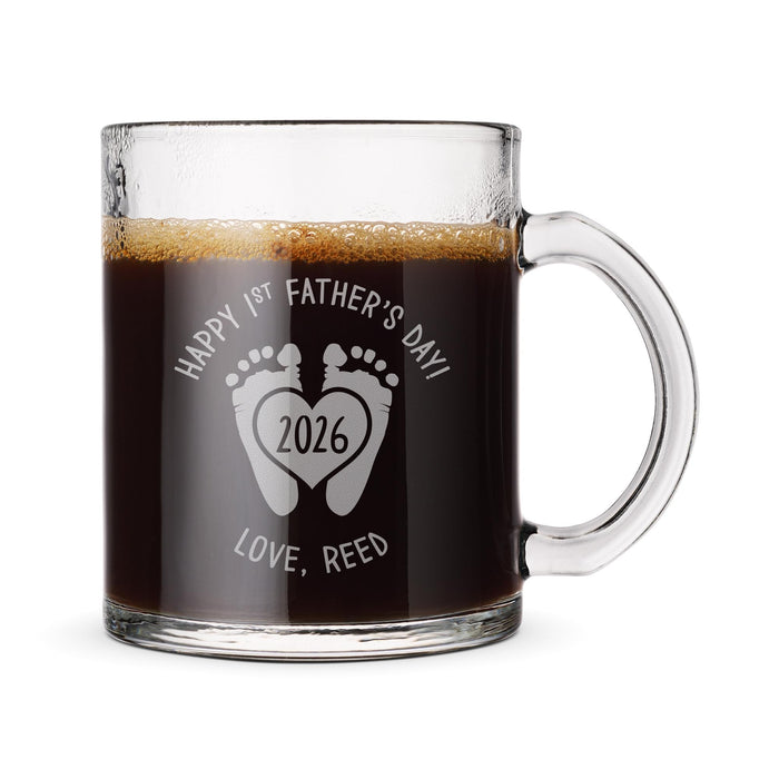 A glass mug filled with a dark liquid, likely coffee or tea, with foam on top. The mug has a handle on the right side and an engraved message on it that reads "Happy 1st Father's Day! Love, Reed" along with a graphic of a heart and the year "2026".