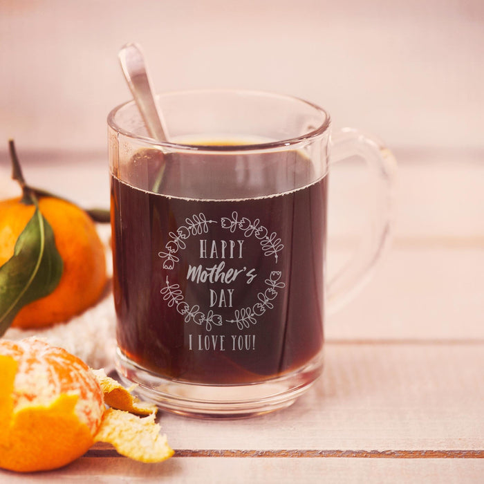 A glass mug filled with a dark liquid, likely coffee, sits on a wooden surface. The mug has a message engraved on it that reads "Happy Mother's Day I love you!"
