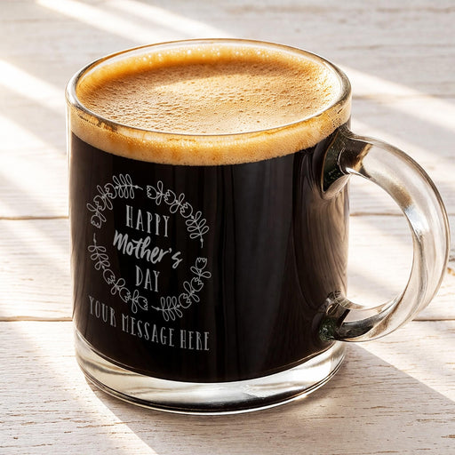 A glass mug filled with a dark liquid, likely coffee, with a frothy top. The mug has a message engraved on it that says "Happy Mother's Day" and "Your Message Here".