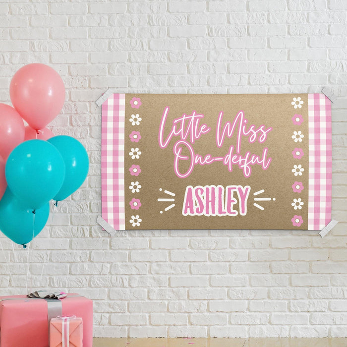 Personalized pink first birthday banner hanging on a white brick wall featuring floral accents and custom printed name design.