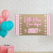 Personalized pink first birthday banner hanging on a white brick wall featuring floral accents and custom printed name design.