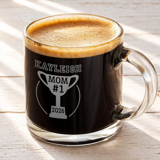 A glass mug filled with a dark liquid, likely coffee, with a foam top. The mug has a personalized message engraved on it, which reads "Kayleigh #1 2026".
