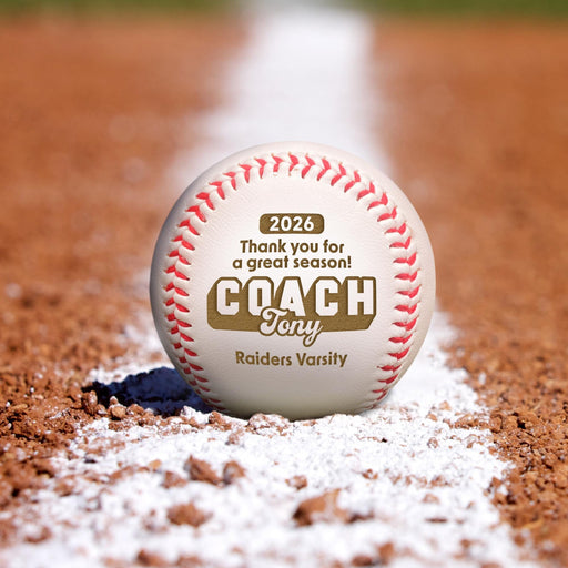 A personalized baseball with the text "2026 Thank you for a great season! COACH Tony Raiders Varsity" printed on it, resting on a baseball field with a white line on the dirt.