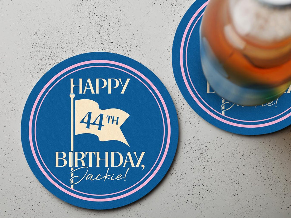 Custom round pulpboard coasters are shown sitting on a light gray stone background with a beer glass on top of one. The custom coasters feature design printed with blue background, pink circular border, and a beige illustrated golf flag.