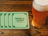 Custom square pulpboard coasters are shown sitting on a wooden table with a pint of beer beside them. The custom coasters feature a green golf bar design with interlocking golf clubs used as a border. The bar name and location can be customized.