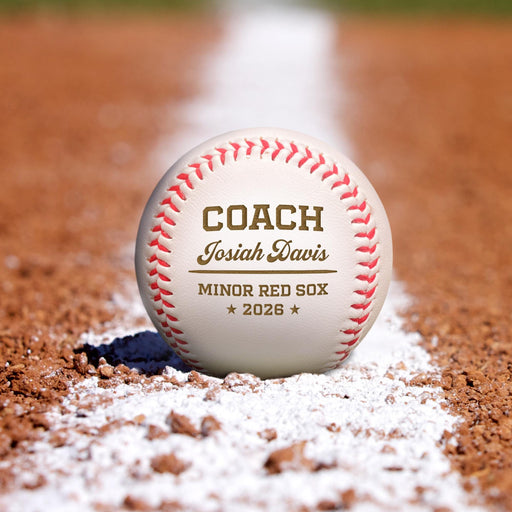 A personalized baseball with the text "COACH Josiah Davis MINOR RED SOX 2026" on it, resting on a baseball field with a white line on the dirt.