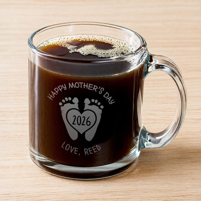 A glass mug filled with a dark liquid, likely coffee, with foam on top. The mug has a personalized message engraved on it, which reads "Happy Mother's Day 2026 Love, Reed".
