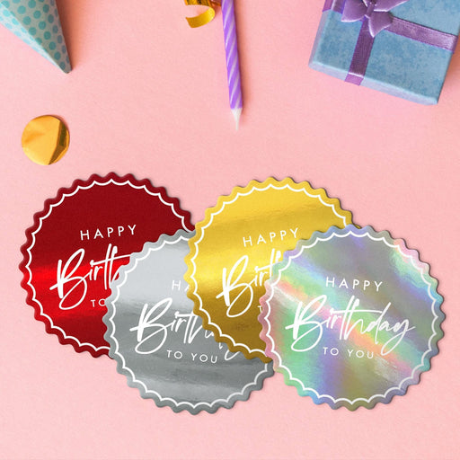 Four birthday gift tags in metallic gold, metallic silver, glitter red, and glitter rainbow lying beside colorful gifts and party decorations.