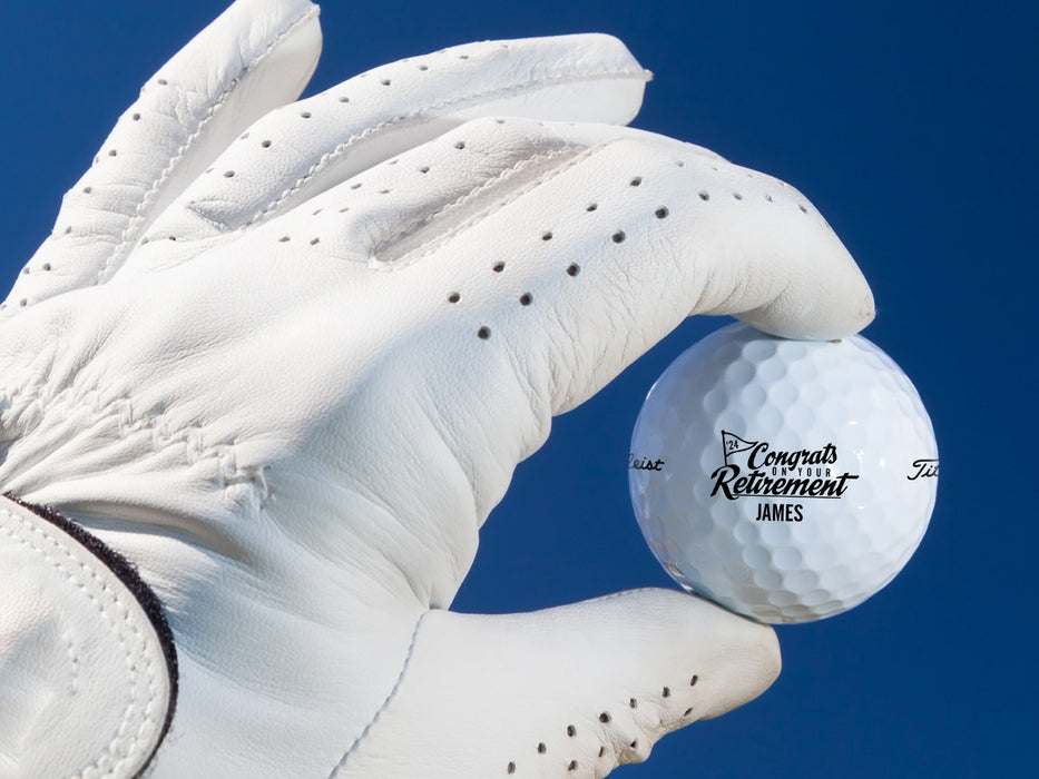 White gloved hand holding single white Titleist golf ball with Congrats On Your Retirement design in front of dark blue background