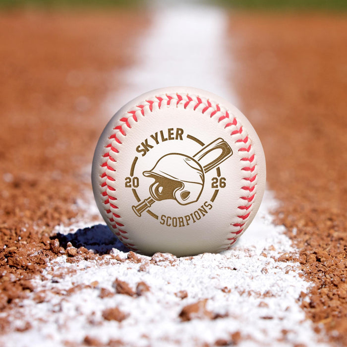 A baseball with the name "SKYLER", the year "2026" engraved on it with a baseball helmet and bat, sitting on a baseball field with a red dirt surface and a white line.