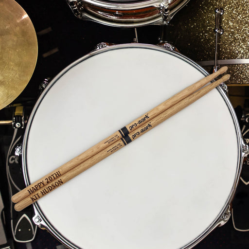 A pair of wooden drumsticks rests on a white drum, with a gold drum visible in the background.