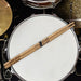A pair of wooden drumsticks rests on a white drum, with a gold drum visible in the background.