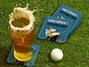 Custom square pulpboard coasters are shown sitting on a grassy, green golf course next to a pint of beer and a white golf ball. The pint of beer has a golf ball splashing into it and has a coaster underneath it. The custom coasters feature a 19th hole bar design with an illustration of a golf ball splashing into a beer mug and a blue background.