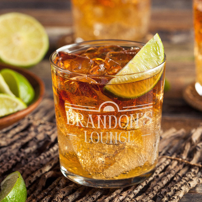 Personalized whiskey glass displayed with whiskey, ice, and fresh lime on a rustic wooden surface. The “Brandon’s Lounge” engraving is clearly visible, showing how the finished custom rocks glass looks in a stylish home bar setting.