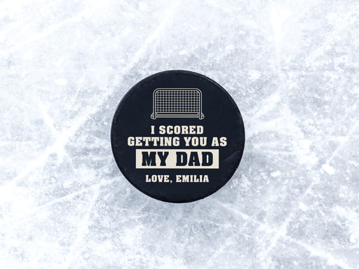 hockey puck on top of ice with white I scored you as my dad design with the name Emilia printed on it