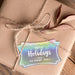 Personalized slim rectangle Christmas gift tag placed on a kraft paper wrapped gift tied with natural twine and dried leaves.