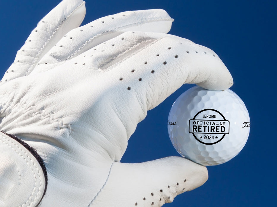 White gloved hand holding single white Titleist golf ball with Officially Retired design in front of dark blue background