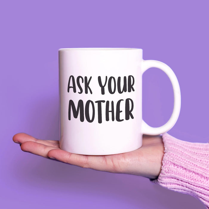 White ceramic mug with the phrase “Ask Your Mother” displayed on an open hand against a soft purple background. A clean product-focused image highlighting the bold black design.