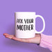 White ceramic mug with the phrase “Ask Your Mother” displayed on an open hand against a soft purple background. A clean product-focused image highlighting the bold black design.