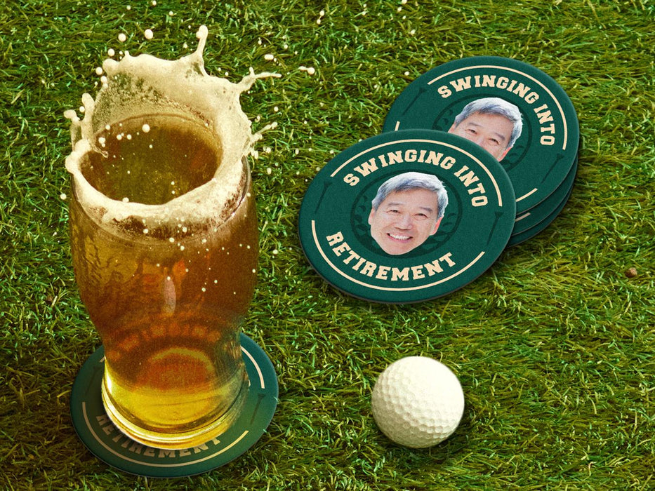 Custom square pulpboard coasters are shown sitting on a grassy, green golf course next to a pint of beer and a white golf ball. The pint of beer has a golf ball splashing into it and has a coaster underneath it. The custom coasters feature design printed with playful &quot;Swinging Into Retirement&quot; text, golf imagery, and custom photo.