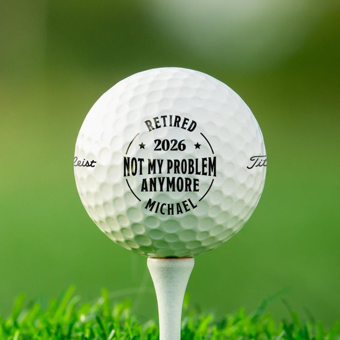 Personalized retirement golf ball featuring the "Not My Problem Anymore" design with a custom name and retirement year. Displayed on a golf tee in grass.