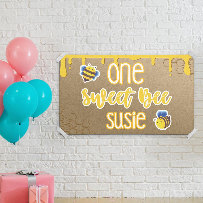 Personalized bee birthday banner hanging on a white brick wall featuring honeycomb accents and custom printed name design.